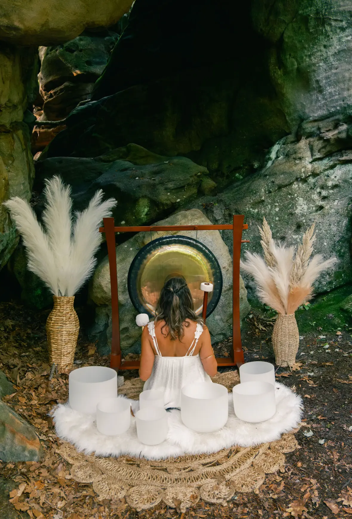 Practitioner seated before a gong and crystal singing bowls in a mossy rock grotto